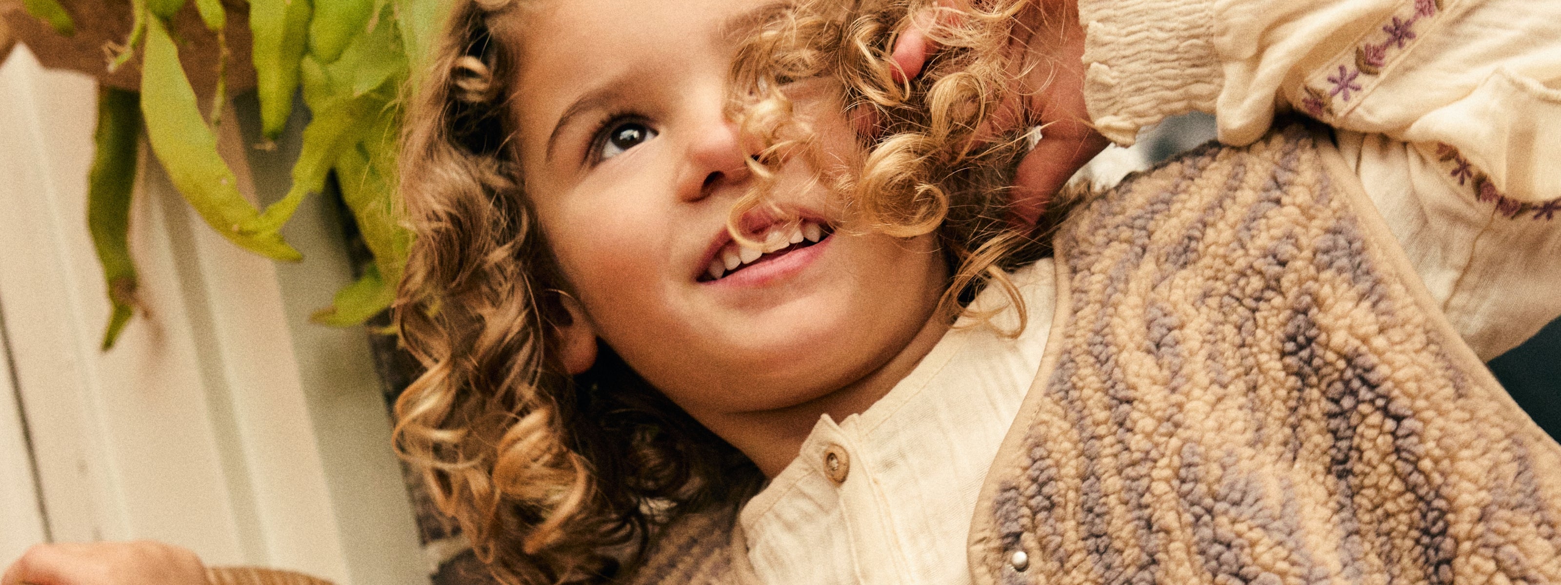 A close-up lifestyle shot of a smiling young girl with curly light brown hair, wearing a cream button-up shirt and a textured brown sherpa vest, with a hand adjusting her hair.