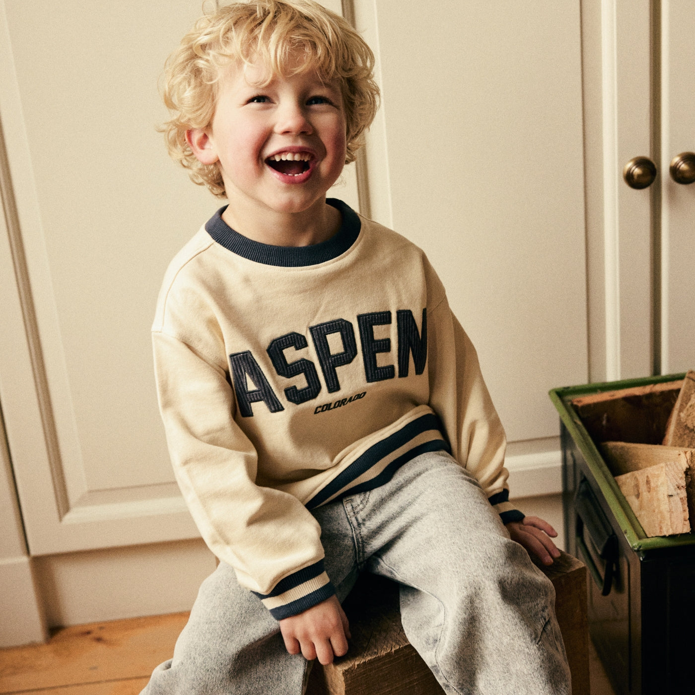 A young child with curly blonde hair sits on a wooden crate in a room, wearing a cream-colored long-sleeve sweatshirt with 'ASPEN COLORADO' printed in navy blue across the chest, paired with gray sweatpants.