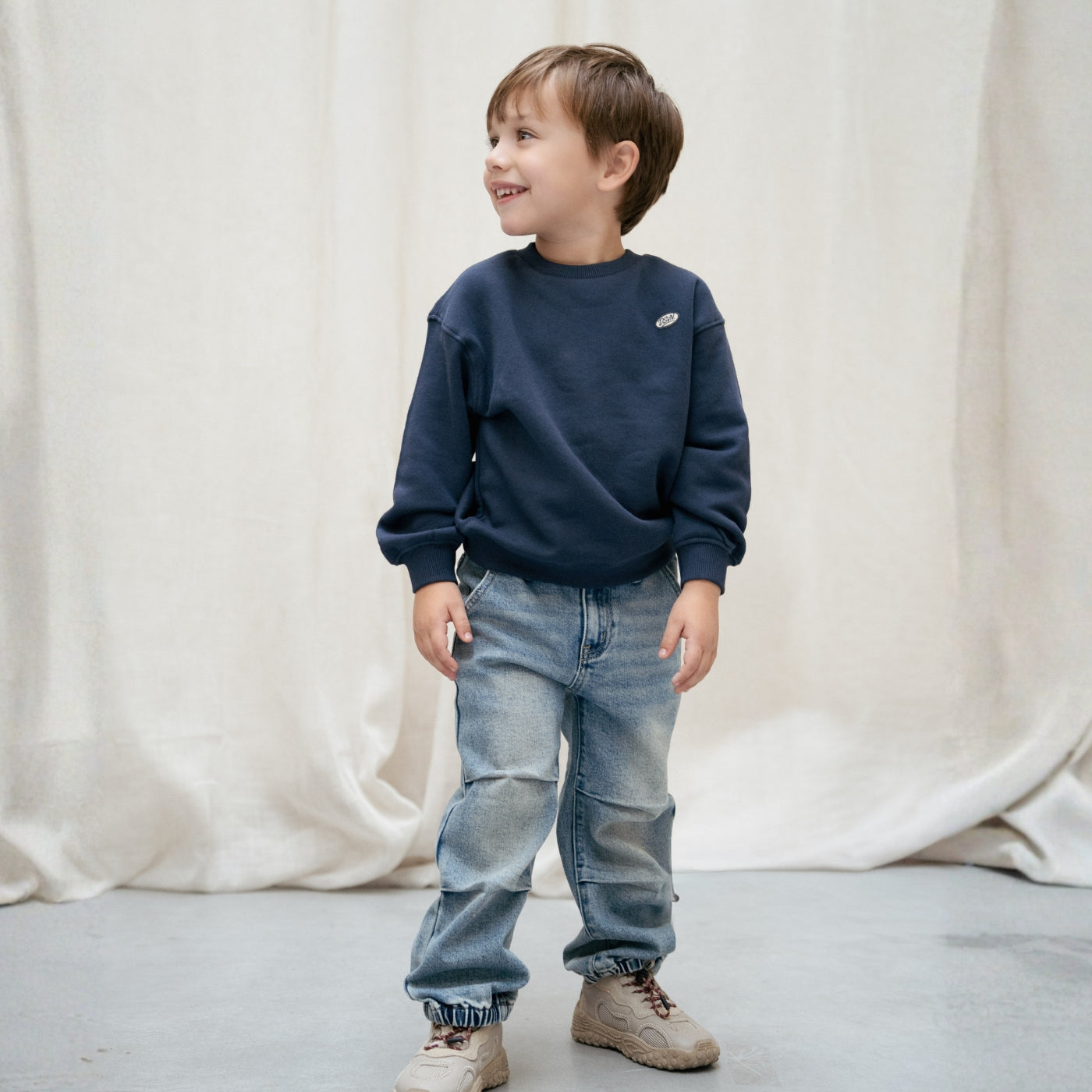 Jongen in een donkerblauwe sweater en lichtblauwe jeans uit de Daily7 Kids collectie.