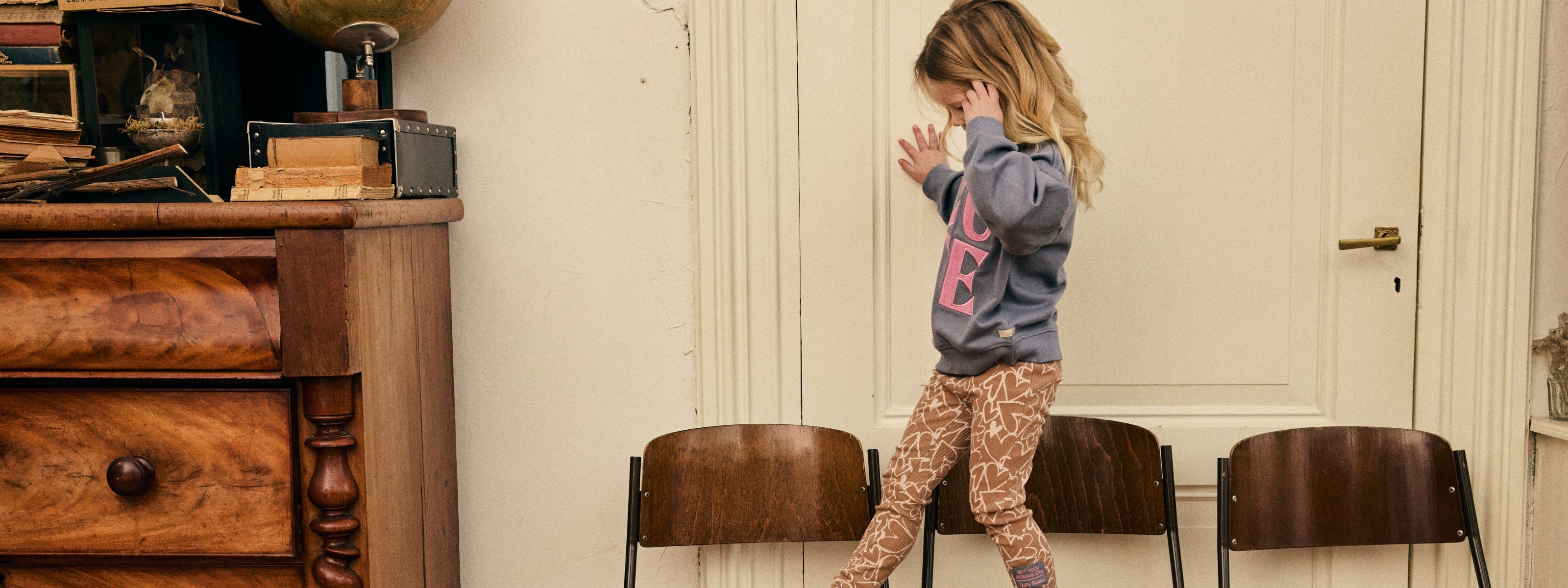 Meisje draagt een sweater met roze artwork en een broek met all-over print uit de Daily7 Kids collectie in een warme, nostalgische setting.