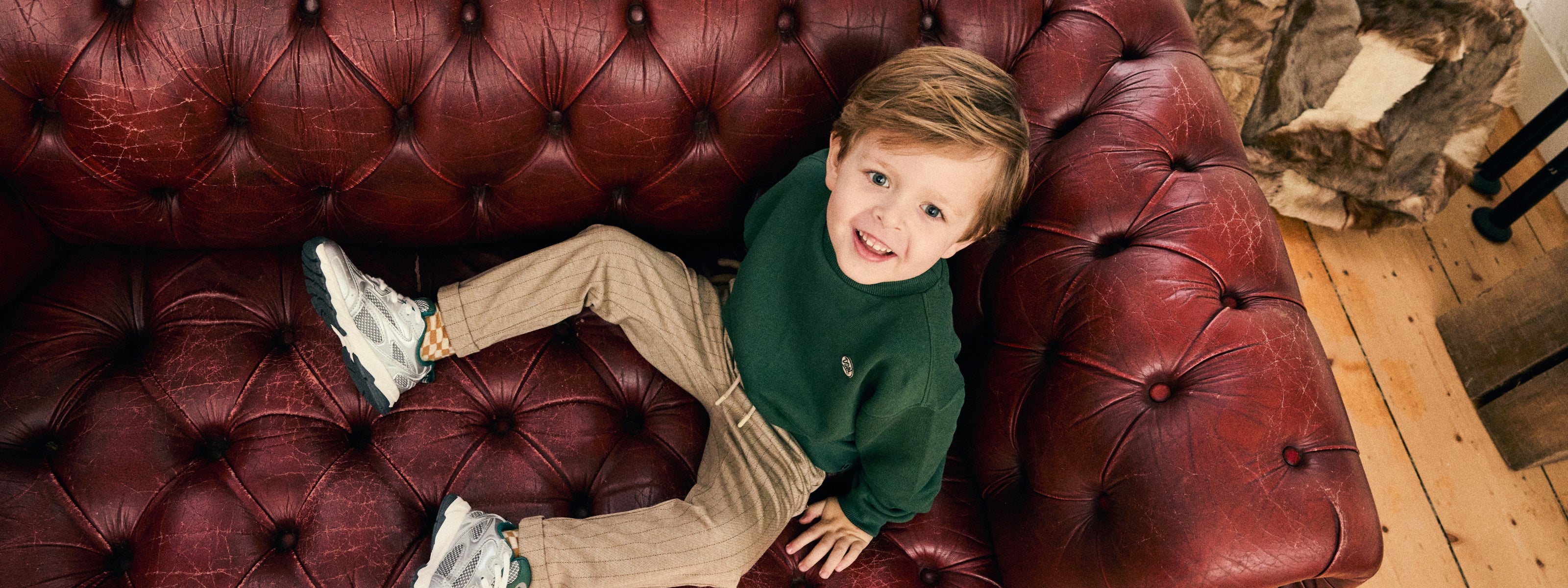 Jongen draagt een donkergroene sweater en een beige broek uit de Daily7 Kids collectie op een vintage leren bank.