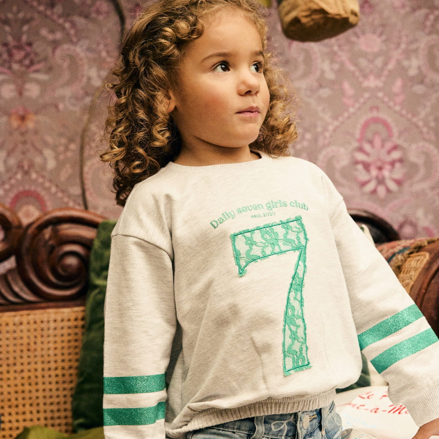 Meisje draagt een Daily7 Kids sweater met groen kanten cijfer 7 in een warme, vintage geïnspireerde setting.