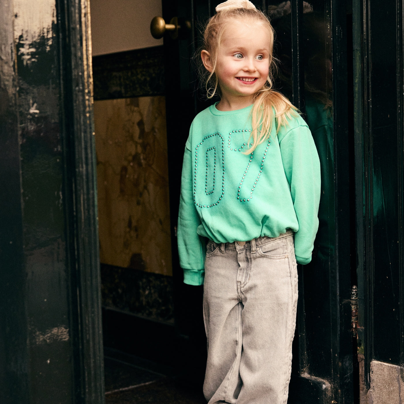 Meisje draagt een mintgroene sweater en een grijze jeans uit de Daily7 Kids collectie in een nostalgisch interieur.