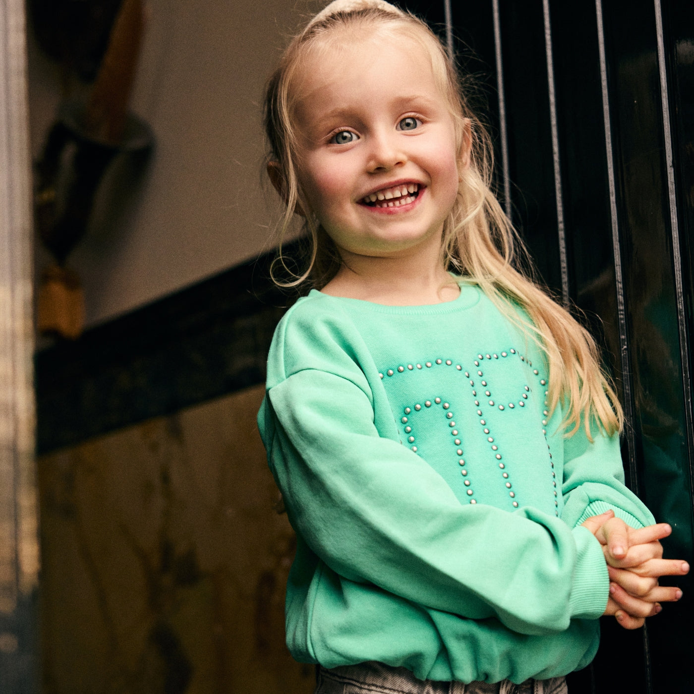 Meisje draagt een mintgroene sweater met studs uit de Daily7 Kids collectie bij een karakteristieke deuropening