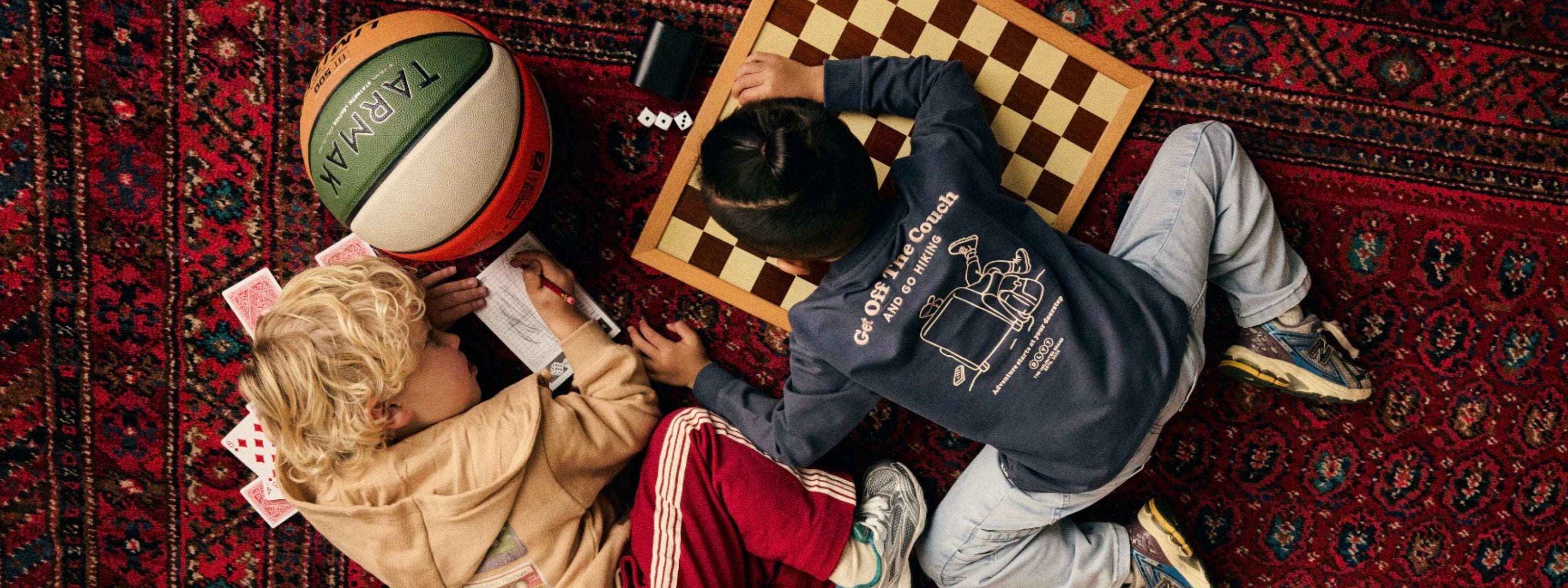 Two children playing on a red patterned rug with a basketball, chessboard, dice, and cards in a cozy indoor setting.