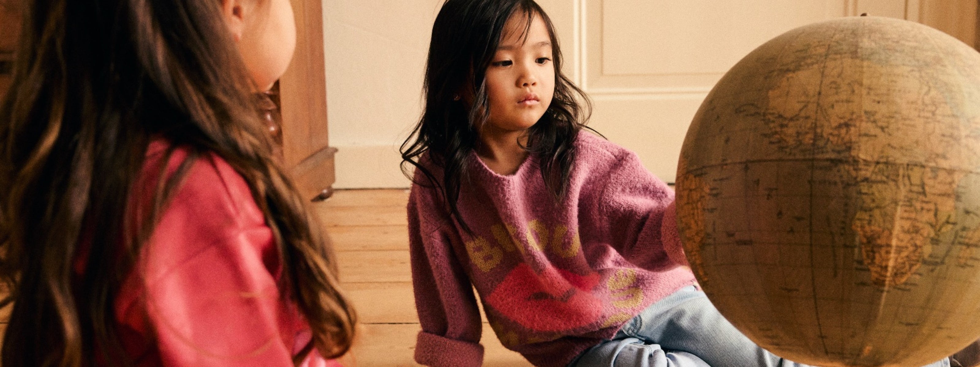 A young girl in a pink knit sweater sits on a wooden floor next to a large vintage globe, with another child partially visible in the foreground in a warm indoor setting.