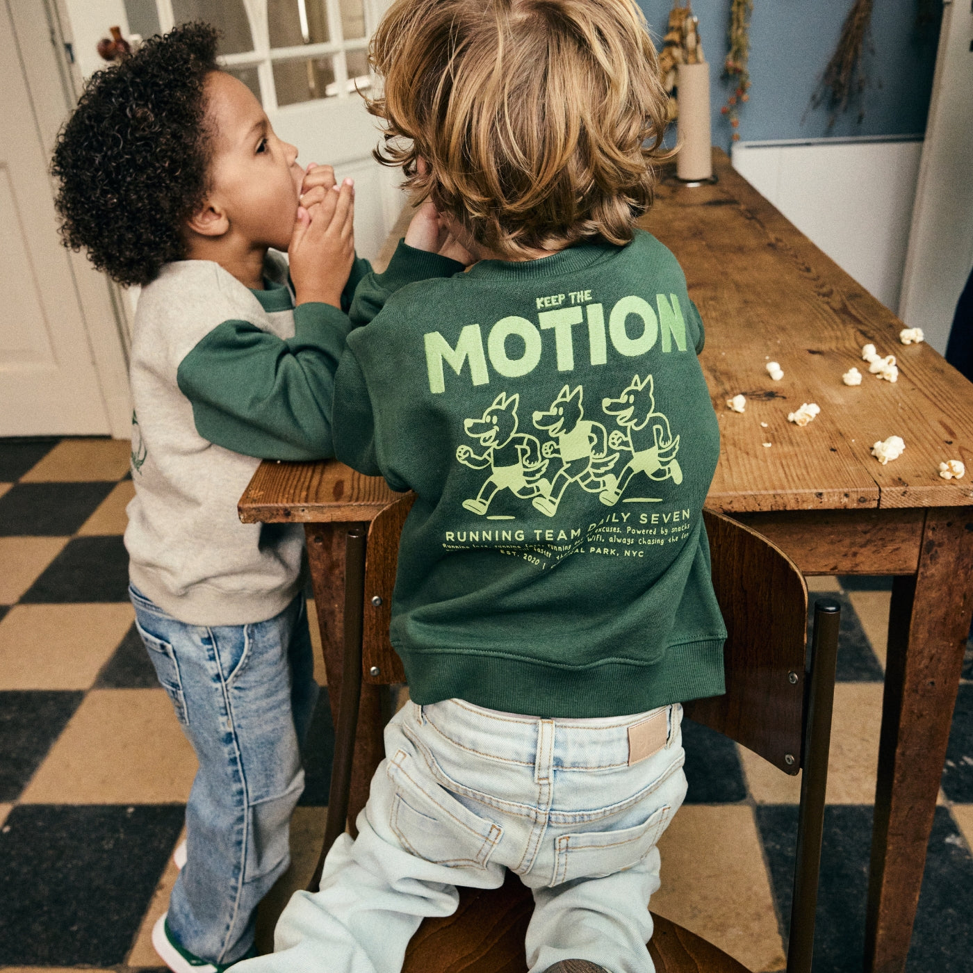Twee jongens dragen sweaters met artwork en jeans uit de Daily7 Kids collectie in een sfeervol interieur.