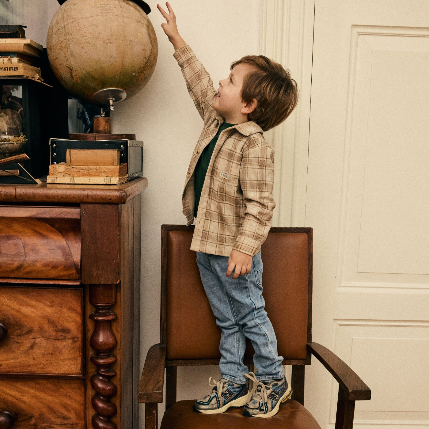Jongen draagt een geruit overshirt en een lichtblauwe jeans uit de Daily7 Kids collectie bij een vintage wereldbol.