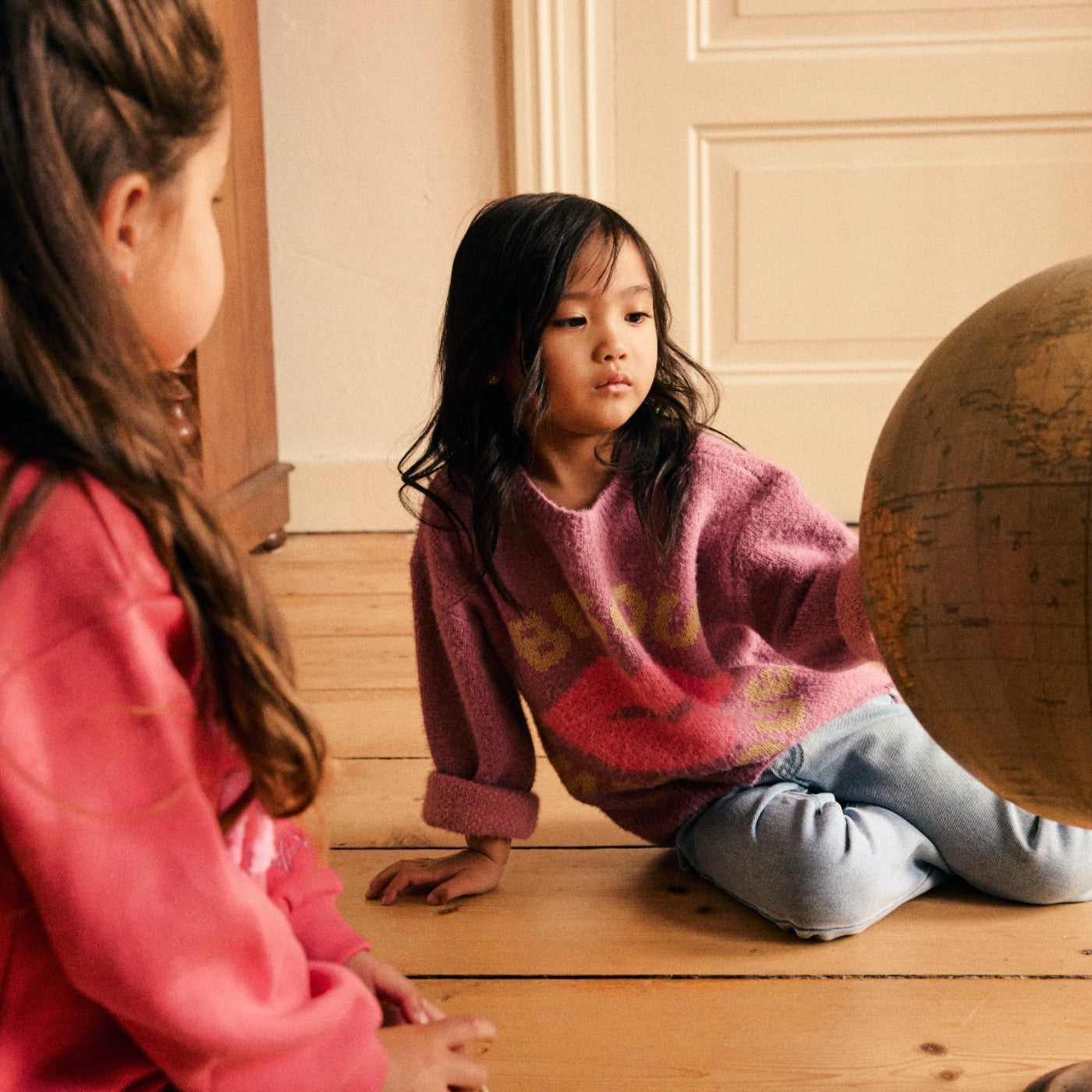 A young girl sits on a wooden floor wearing a fuzzy pink sweater and light blue jeans, looking at another child in a coral-pink sweatshirt. A large vintage globe is partially visible on the right, and a cream-colored paneled door is in the background.