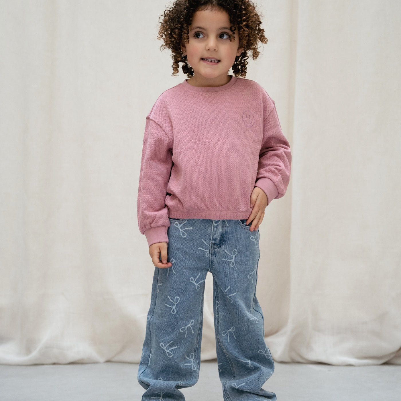 A young child with curly brown hair stands against a draped cream fabric backdrop, wearing a dusty pink long-sleeve sweatshirt and light blue jeans with a white bow print.