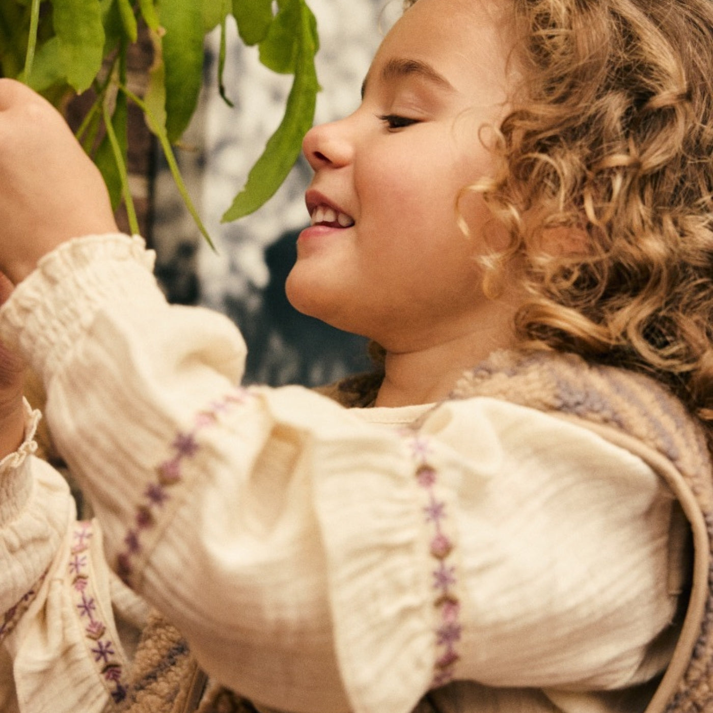 A close-up lifestyle shot of a young child with curly light brown hair, wearing a cream-colored long-sleeve blouse with purple embroidered details and a brown vest, looking up at green leaves.