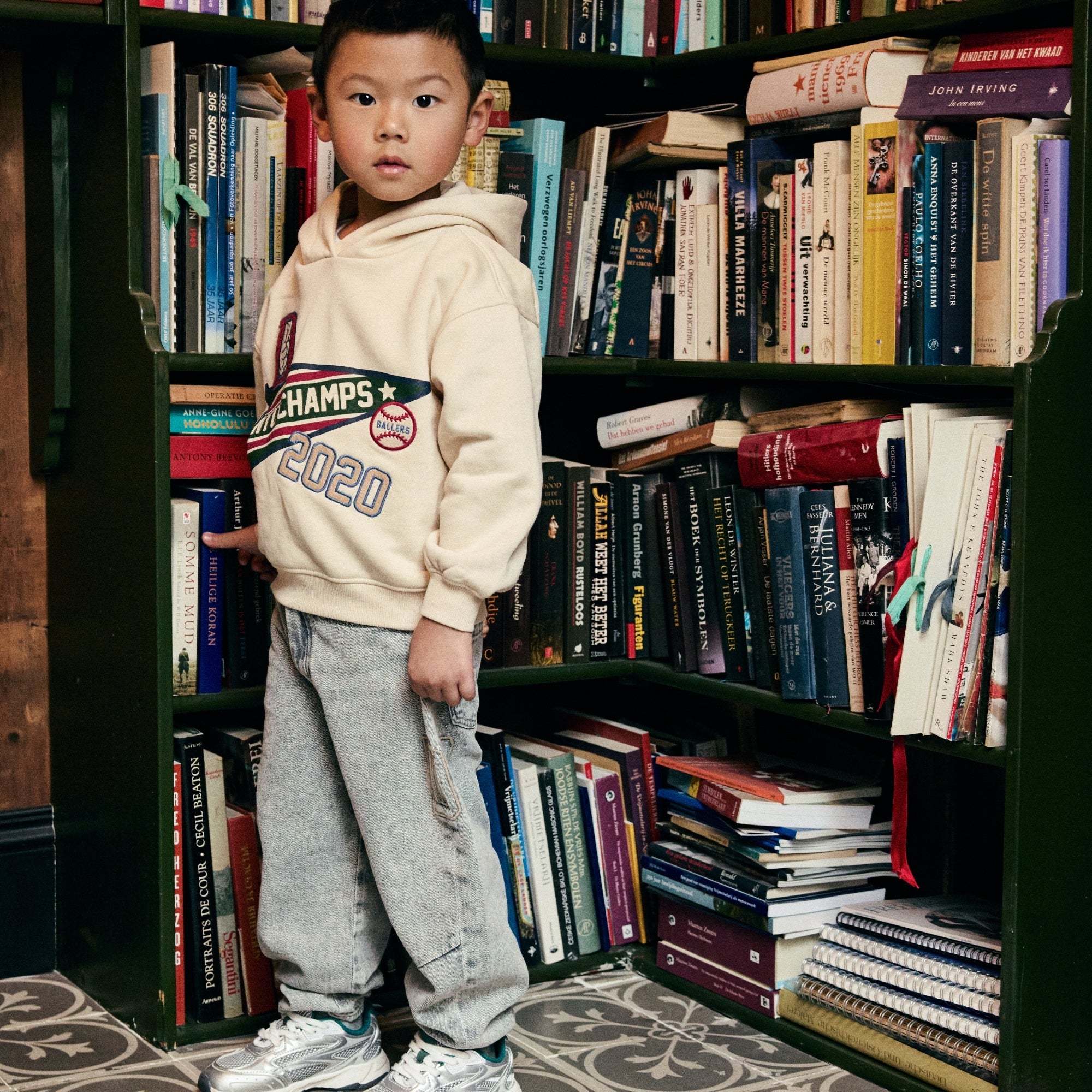 Jongen draagt een hoodie en een lichtblauwe jeans uit de Daily7 Kids collectie voor een gevulde boekenkast.