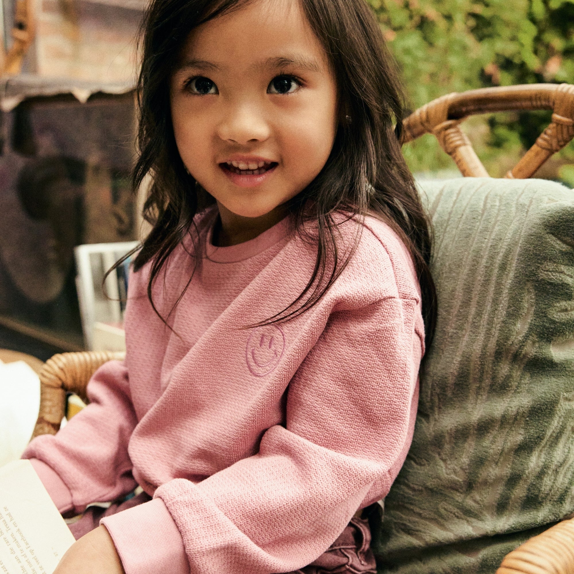 Meisje draagt een roze sweater met smiley-detail uit de Daily7 Kids collectie in een sfeervolle setting.