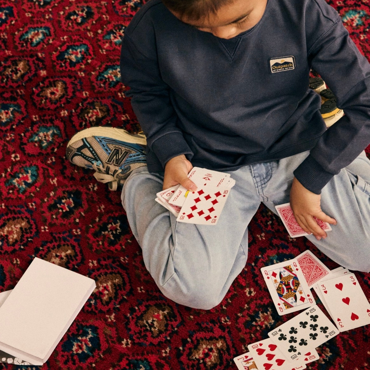 A child sitting on a red patterned rug playing with playing cards. The child is wearing a navy blue Patagonia sweatshirt, light blue jeans, and sneakers.