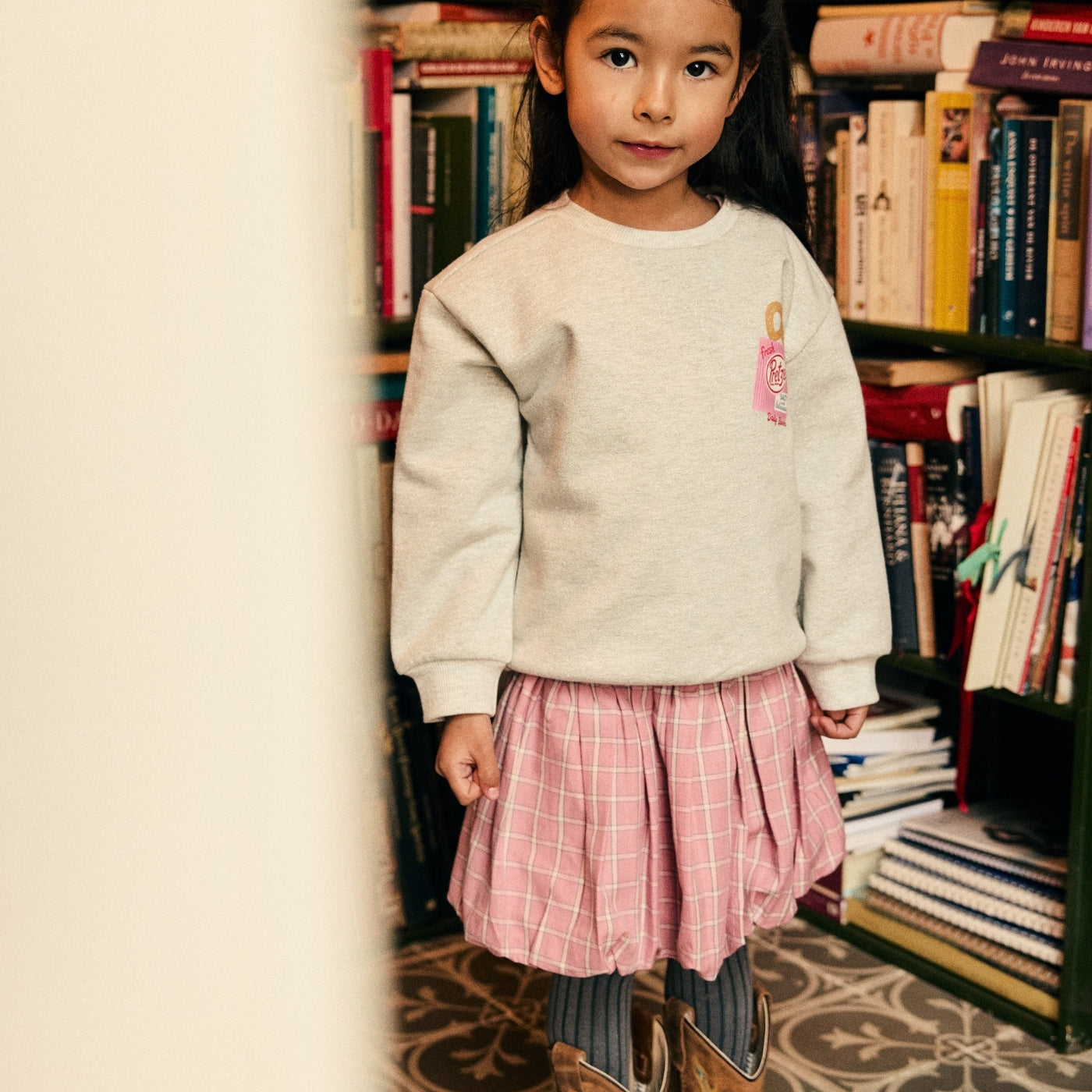 A young girl stands in a book-filled room wearing a light gray crewneck sweatshirt, a pink plaid pleated skirt, striped leggings, and brown cowboy boots.